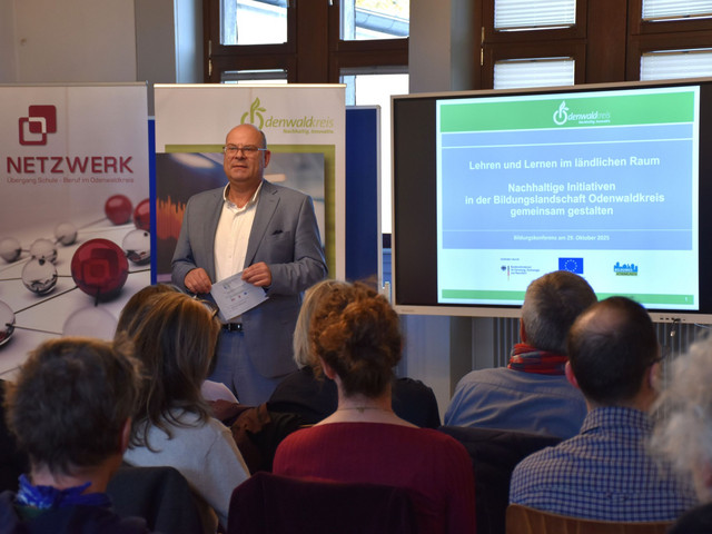 Das Bild zeigt den Ersten Kreisbeigeordneten Oliver Grobeis, der vor einem Publikum steht und spricht. Im Hintergrund sind Banner und ein Bildschirm mit der Aufschrift "Lehren und Lernen im ländlichen Raum" zu sehen. Bei der Veranstaltung geht es um Bildung und nachhaltige Initiativen im Odenwaldkreis.