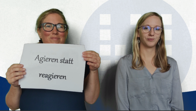 Zwei jüngere Frauen blicken in die Kamera. Eine von ihnen hält ein Schild mit der Aufschrift "Agieren statt Reagieren" hoch.