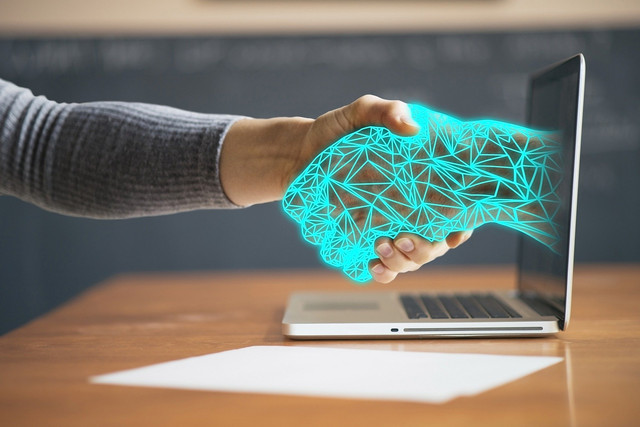 Ein Unterarm mit einer Hand tritt aus einem Notebook-Bildschirm hervor und ergreift eine physische Hand. 