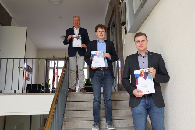 Drei Männer verschiedenen Alters stehen auf einer Treppe und halten ein Druckerzeugnis hoch.