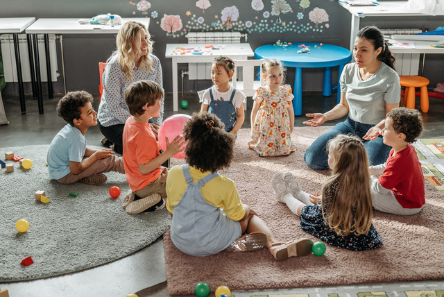 Mehrere jüngere Kinder sitzen im Kreis um zwei Betreuerinnen herum.