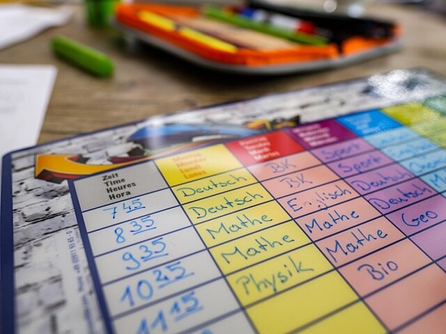Ein Stundenplan mit den Fächern Deutsch, Mathe, Physik, Bio, Englisch und Sport liegt auf einem Holztisch. Im Hintergrund ist ein Schulmäppchen mit Buntstiften zu sehen, die auf einen Schultag vorbereitet. Der Stundenplan symbolisiert Struktur und Organisation im Schulalltag eines Kindes