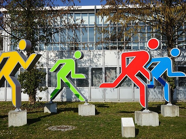 Auf dem Bild sind vier Skulpturen zusehen. Sie stellen vier stilisierte Figuren dar, die in leuchtenden Farben – Gelb, Grün, Rot und Blau – gehalten sind und sich in Lauf- oder Sprungbewegungen befinden. Sie sind auf Betonfundamenten im Freien aufgestellt, vor einer modernen Gebäudefassade mit großen Fenstern. Die Figuren scheinen Dynamik und Fortschritt zu symbolisieren, möglicherweise im Kontext von Sport, Wettbewerb oder auch dem Start in neue Unternehmungen. Die bunten Farben und die energischen Posen vermitteln eine positive und motivierende Botschaft.