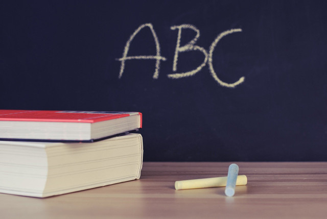 Im Hintergrund steht auf einer Tafel in Kreide geschrieben „ABC“. Davor liegen auf einem Tisch zwei Bücher aufeinander, daneben zwei Stück Kreide in den Farben Hellbau und Hellgelb.