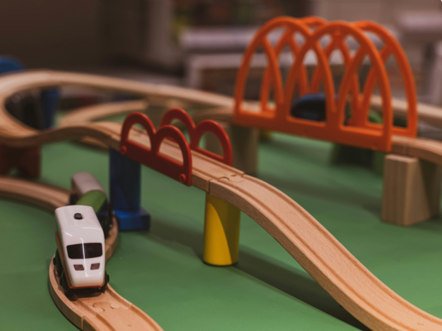 Das Bild zeigt eine Spielzeugeisenbahn aus Holz, die auf einer grünen Oberfläche aufgebaut ist. Die Bahn besteht aus hellbraunen Holzschienen, die sich in Kurven und über Brücken winden. Eine der Brücken ist orangefarben und hat eine bogenförmige Struktur, während eine andere rot ist und von farbigen Säulen gestützt wird. Ein weißer Spielzeugzug mit schwarzen Fenstern und roten Akzenten steht auf den Gleisen. 