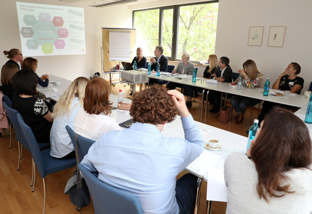 Forumsteilnehmende sitzen in U-Form zusammen und blicken auf die Beamerleinwand im Vordergrund des Seminarraums 4..