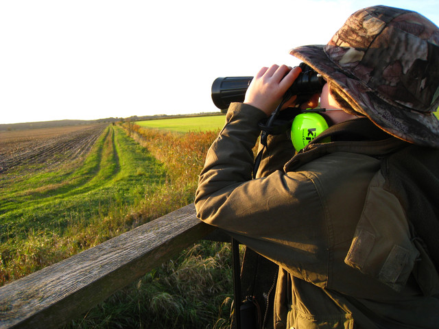 Mensch mit Hut betrachtet eine Ackerlandschaft durch ein Fernglas.