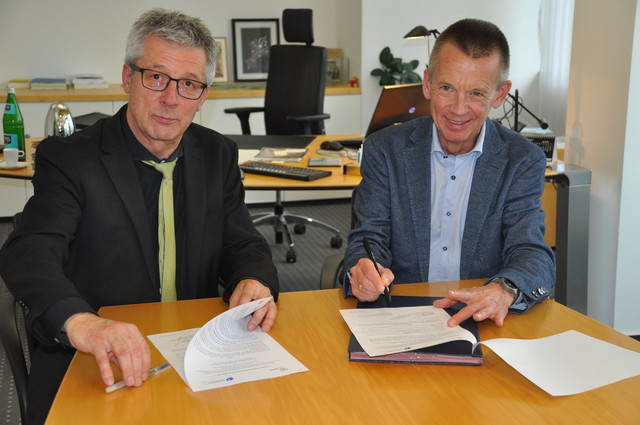 Zwei Männer in Anzügen sitzen am Tisch und signieren eine Kooperationserklärung.