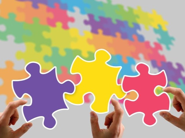 Das Bild zeigt drei farbige Puzzleteile, die von jeweils einer Hand gehalten werden und zusammengefügt werden sollen, vor einem Hintergrund aus vielen bunten Puzzleteilen. Die drei im Vordergrund befindlichen Puzzleteile symbolisieren die Zusammenarbeit, Integration und den gemeinsamen Erfolg. Die Vielfalt der Farben im Hintergrund unterstreicht die Bedeutung von Diversität und Inklusion, um ein gemeinsames Ziel zu erreichen.