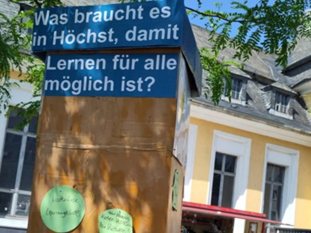 Das Bild zeigt eine Säule als eine Art Umfrage- oder Sammelstelle im öffentlichen Raum, die sich mit der Frage beschäftigt, was in Höchst benötigt wird, damit Lernen für alle möglich ist. An der Säule sind Zettel mit Antworten oder Vorschlägen befestigt.
