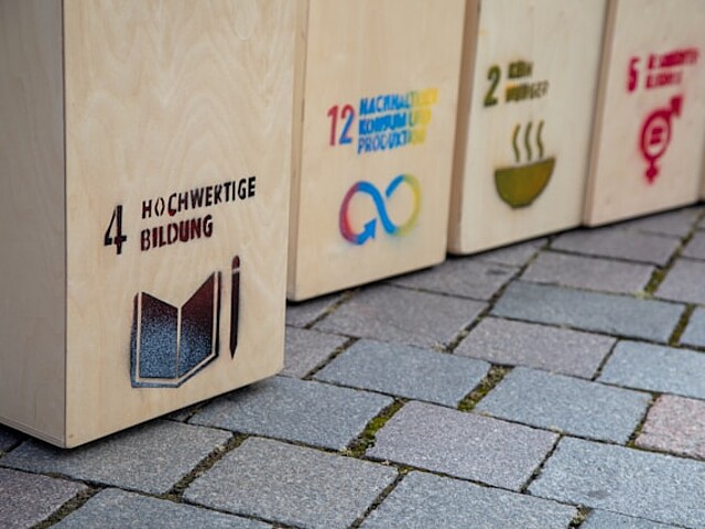 Es stehen mehrere Holzkisten auf einem gepflasterten Boden nebeneinander. Auf der vorderen Kiste ist das Ziel „Hochwertige Bildung“ mit einem Buchsymbol abgebildet. Auf den weiteren Kisten sind Symbole weiterer Ziele für nachhaltige Entwicklung zu sehen, darunter nachhaltiger Konsum und Produktion, Maßnahmen zum Klimaschutz und Geschlechtergleichheit. Die Kisten sollen den Zusammenhang zwischen Bildung für nachhaltige Entwicklung und den globalen Nachhaltigkeitszielen verdeutlichen