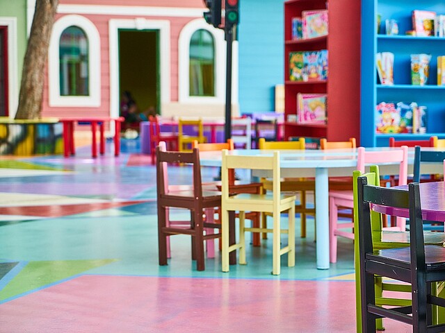 Dieses Bild zeigt einen hellen und farbenfrohen Raum in einem Kindergarten oder einer Kita. Man sieht mehrere kleine Tische und Stühle in leuchtenden Farben wie Pink, Gelb, Grün und Braun, die um die Tische gruppiert sind. Im Hintergrund sind bunte Regale voller Bücher und Spielsachen sowie eine Fassade, die wie ein kleines buntes Haus aussieht, was die spielerische Atmosphäre unterstreicht.