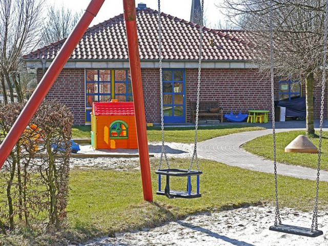 Die Gebäudefassade eines Kindergartens oder einer Kindertagesstätte ist im Hintergrund des Fotos zu sehen. Im Vordergrund befinden sich Spielgeräte für Kinder, darunter eine Schaukel; dazwischen grüne Rasenflächen. Personen sind nicht auf dem Bild sichtbar. 