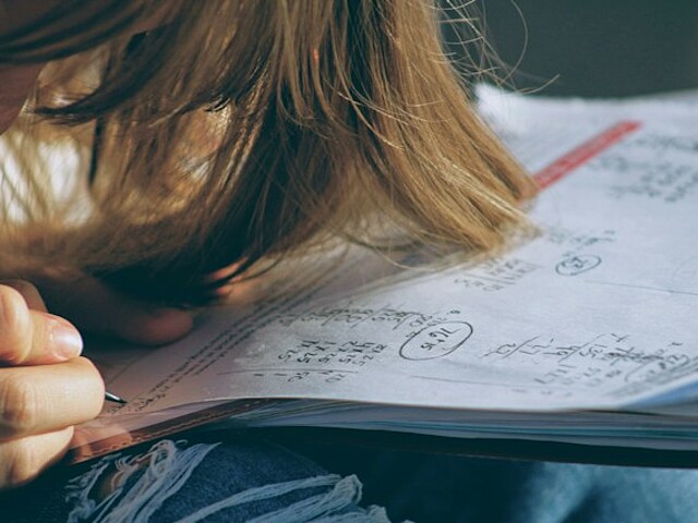 Im Vordergrund sind die langen braunen Haaren einer Person zu sehen, die sich über ein aufgeschlagenes Heft beugt. Die Person schreibt gerade mit der Hand in das Heft, auf dessen Seiten sich handschriftliche Notizen und Rechenaufgaben befinden. Die Aufnahme vermittelt eine konzentrierte Lernsituation.